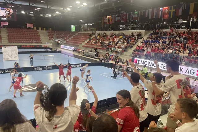 Punaiset ja siniset pelaajat mailoineen salibandyottelussa, katsomo kannustaa